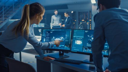 A woman in a high-tech work setting points at a computer screen to show a colleague something. 